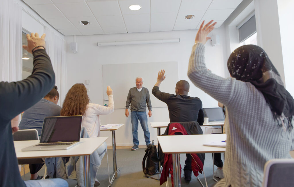 Elever med olika etnicitet räcker upp handen framför läraren under vuxenutbildning / Svenska för invandrare